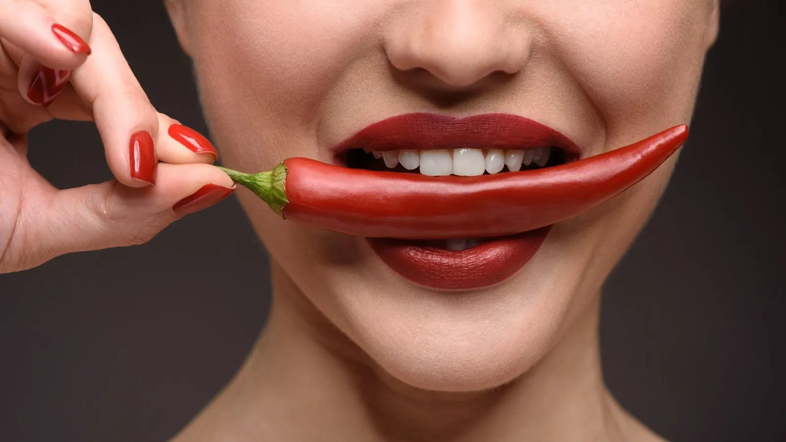 Untere Hälfte des Gesichts einer Frau, die roten Lippenstift trägt und eine Chilischote zwischen den Lippen hält.